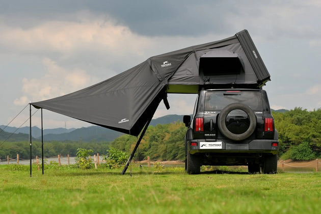 Black rooftop tent attached to a black SUV in a grassy field with mountains in the background.